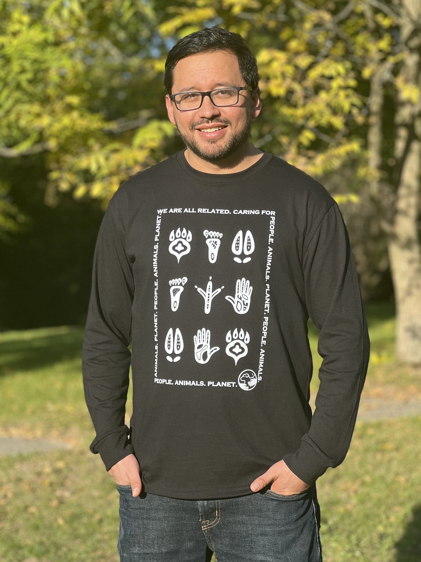 A man wearing a long-sleeved black t-shirt with a white print that is made up of Indigenous-style footprints of animals and humans with the words "We are all related. Caring for People. Animals. Planet." in a rectangular shape around the prints.