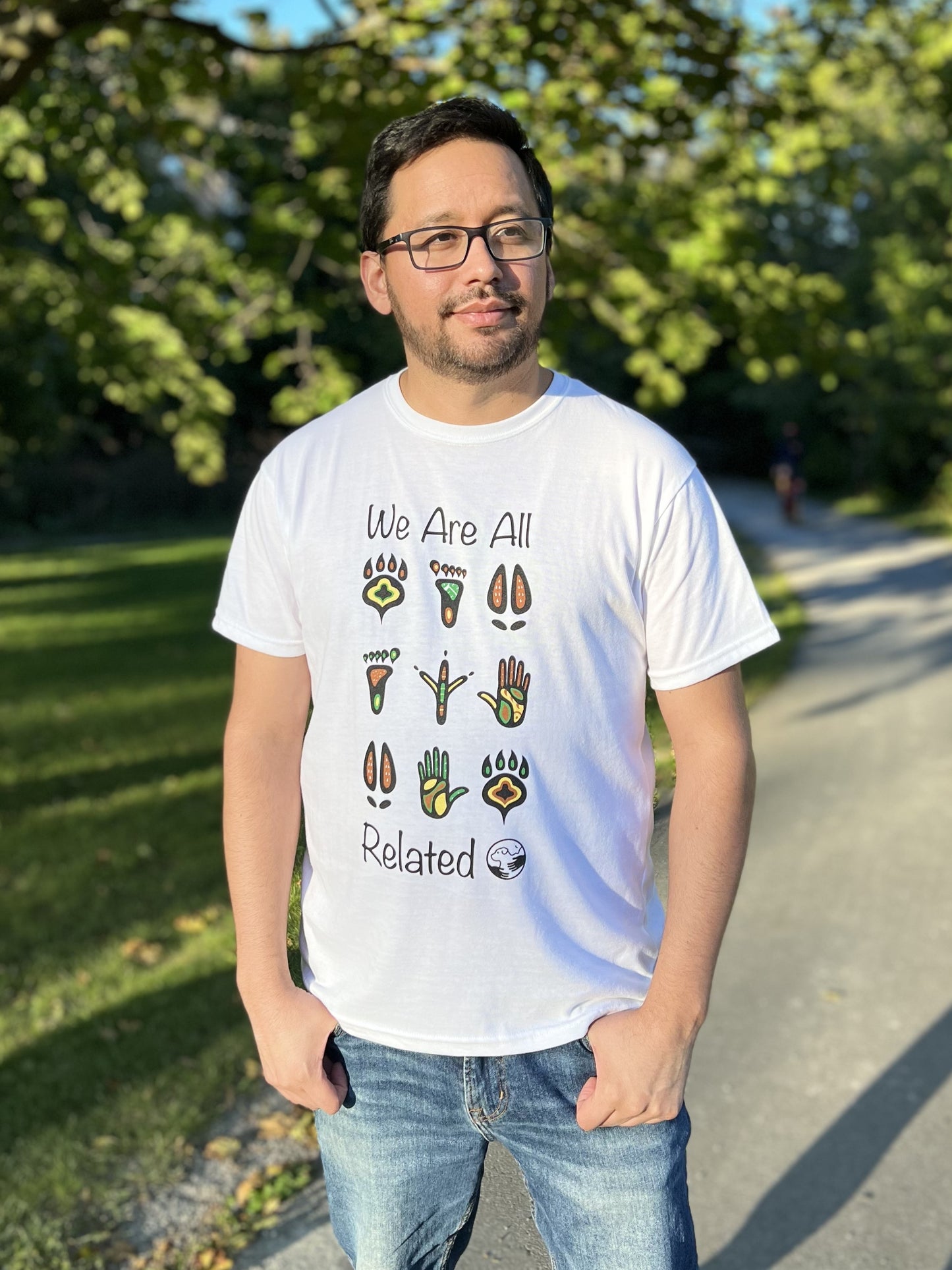 A man wearing a white t-shirt with a mutlti-coloured print of stylized animal and human footprints. The words "We are all" are above the footprints "Related" is below the footprints with the One Health Partners logo beside.