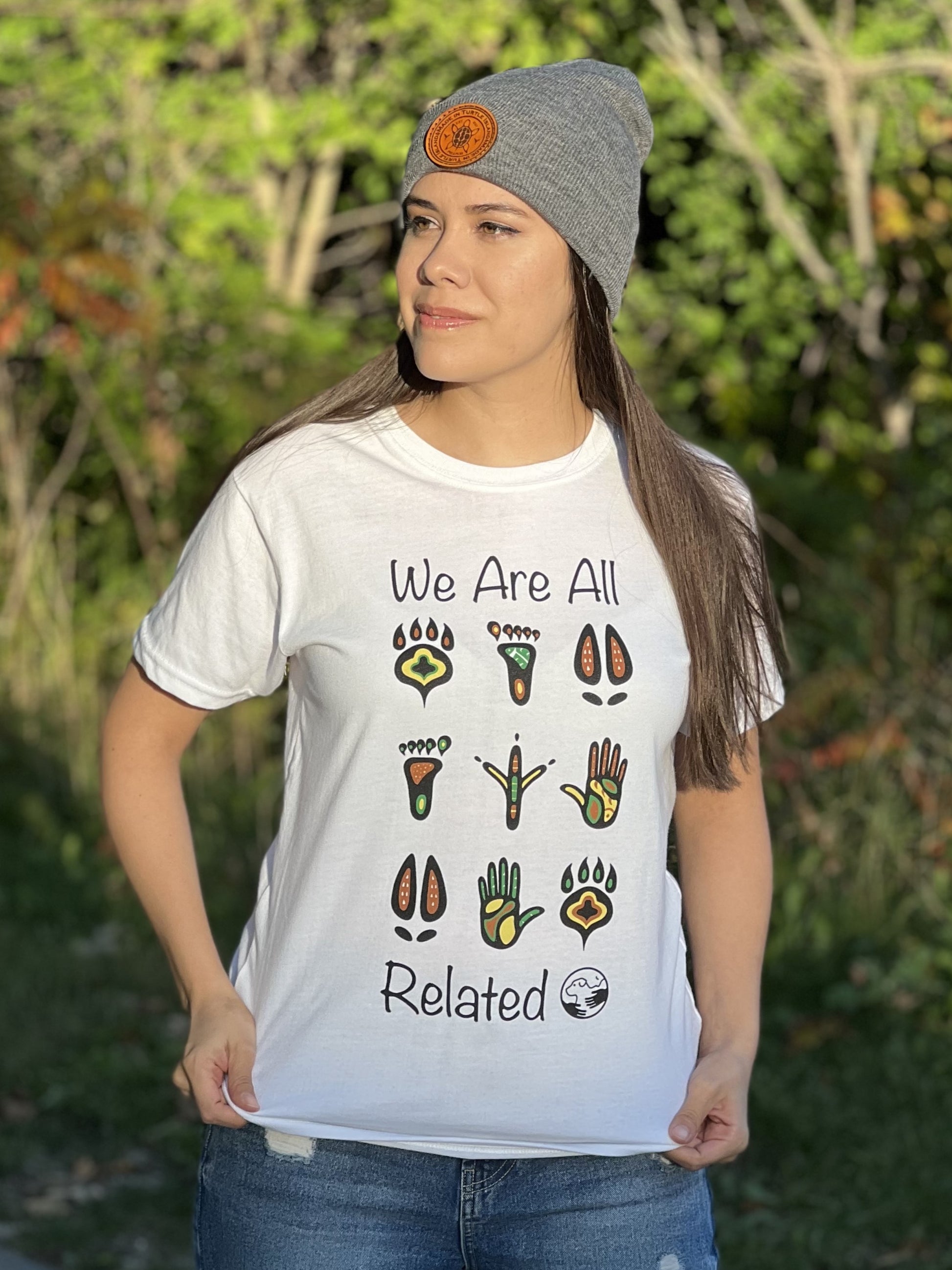 A woman wearing a white t-shirt with a mutlti-coloured print of stylized animal and human footprints. The words "We are all" are above the footprints "Related" is below the footprints with the One Health Partners logo beside. 