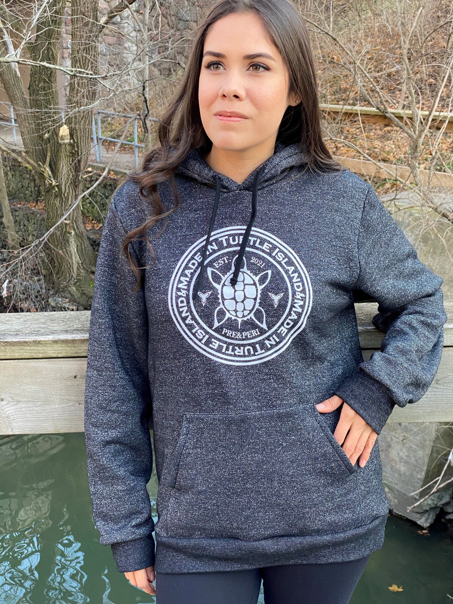 A woman wearing a forest green hoodie with kangaroo pocket. White artwork of a stylized turtle with the words "Made in Turtle Island" around it.