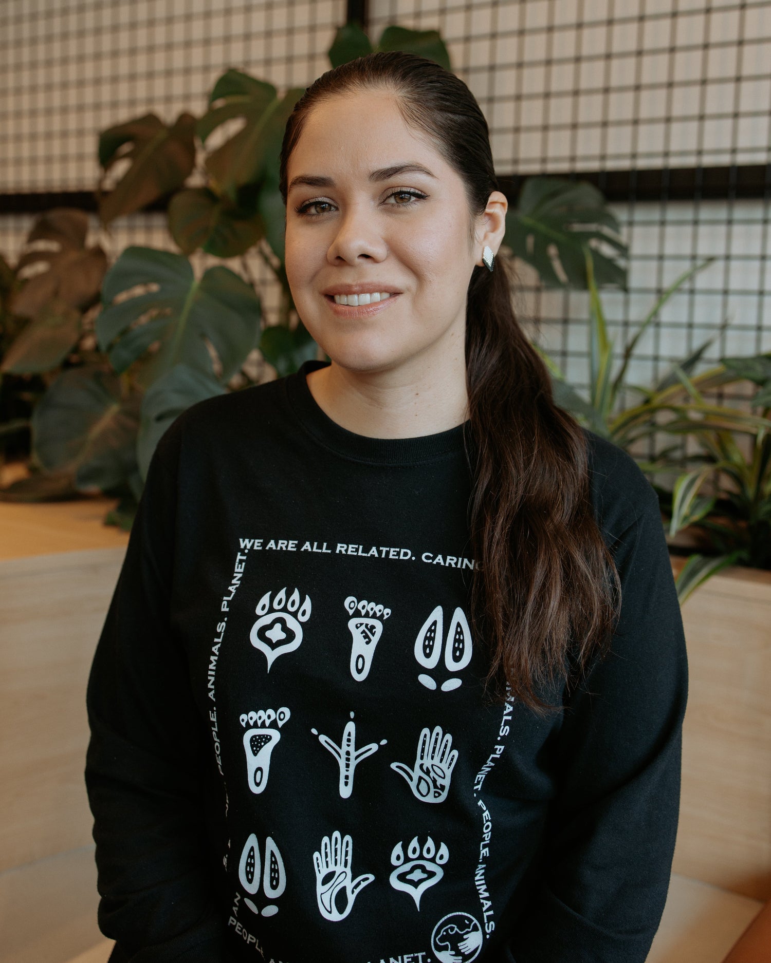 A smiling woman with dark hair tied back is wearing a black crewneck sweatshirt with white animal paw and hand graphics and the text 'WE ARE ALL RELATED' She is photographed indoors in front of a grid-patterned screen and green plants.