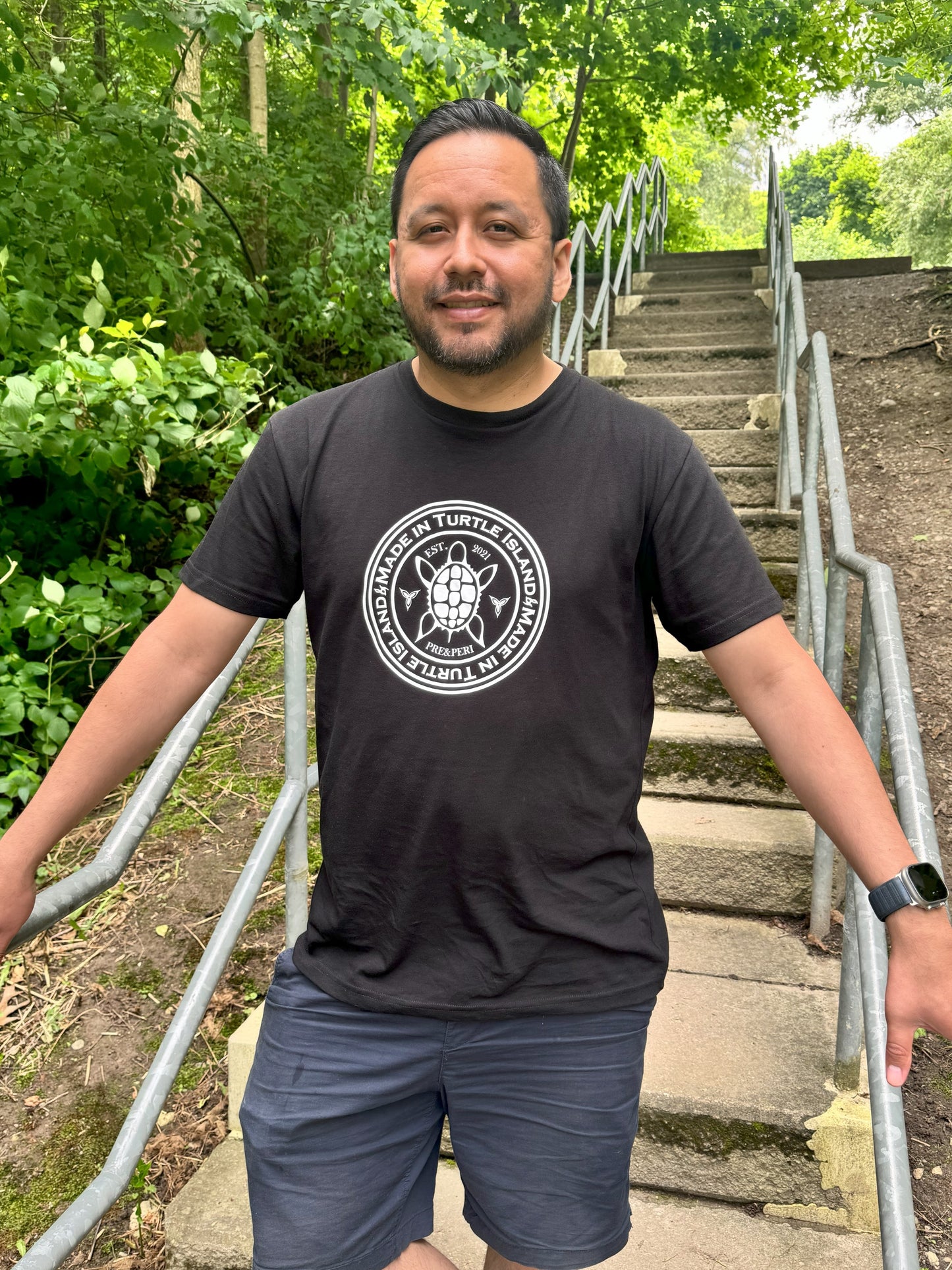 A man standing on outdoor steps wearing a black organic cotton t-shirt with a white print that says "Made in Turtle Island" around a central image of a turtle.