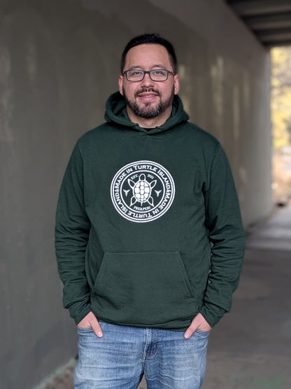 A man wearing a forest green hoodie with kangaroo pocket. White artwork of a stylized turtle with the words "Made in Turtle Island" around it.