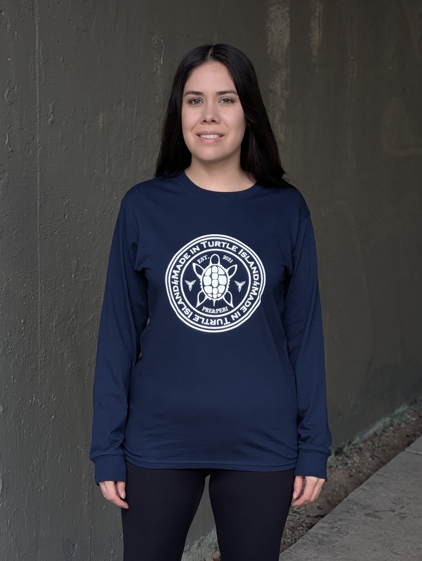 A woman wearing a navy long sleeve t-shirt with a white print of a turtle in the centre of a circle with words "Made in Turtle Island" around it twice