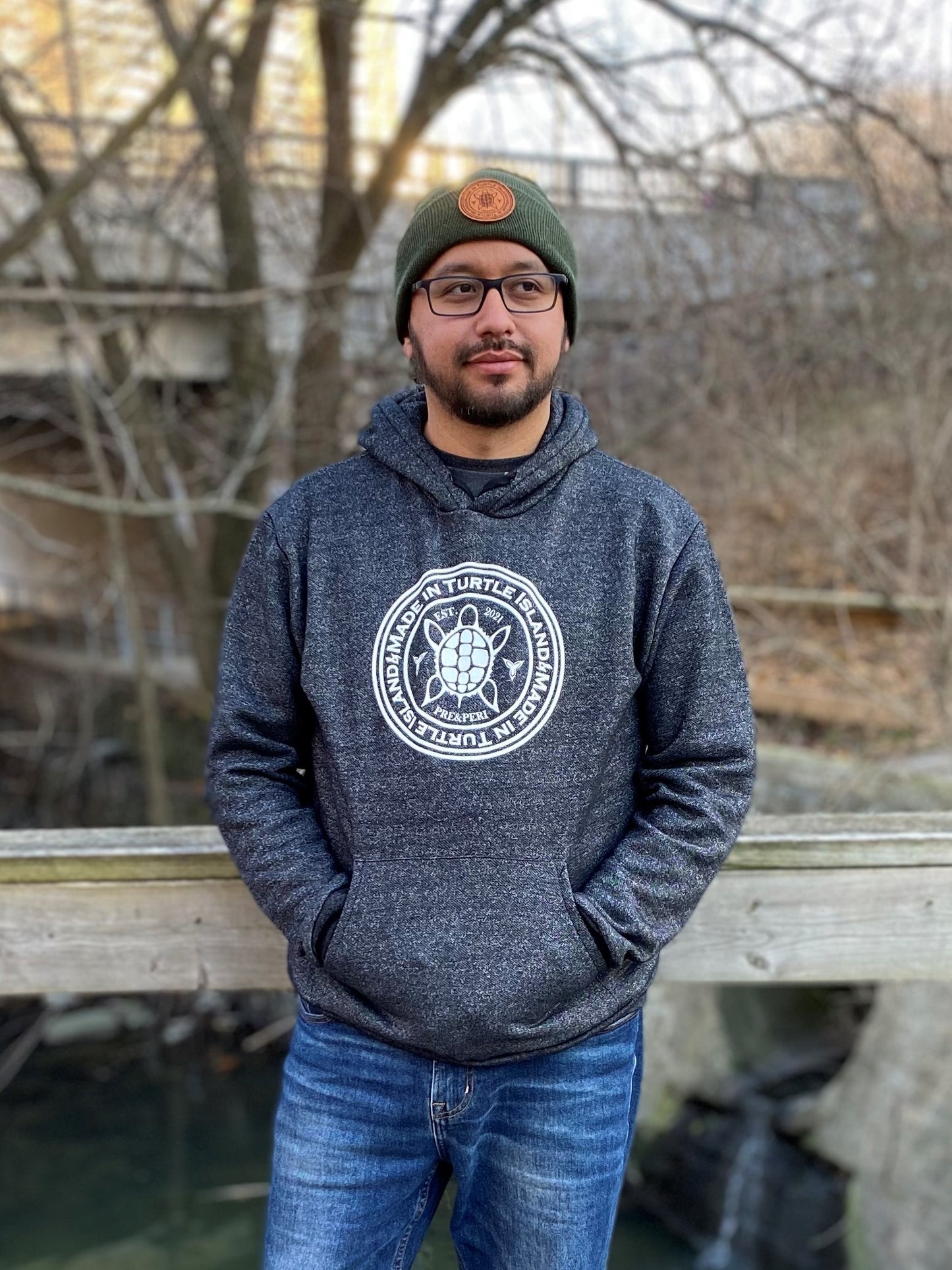 A bearded man wearing glasses, a green toque with tan leather patch, and salt and pepper hoodie with white screen printed graphic featuring a turtle with the words "Made in Turtle Island" around it